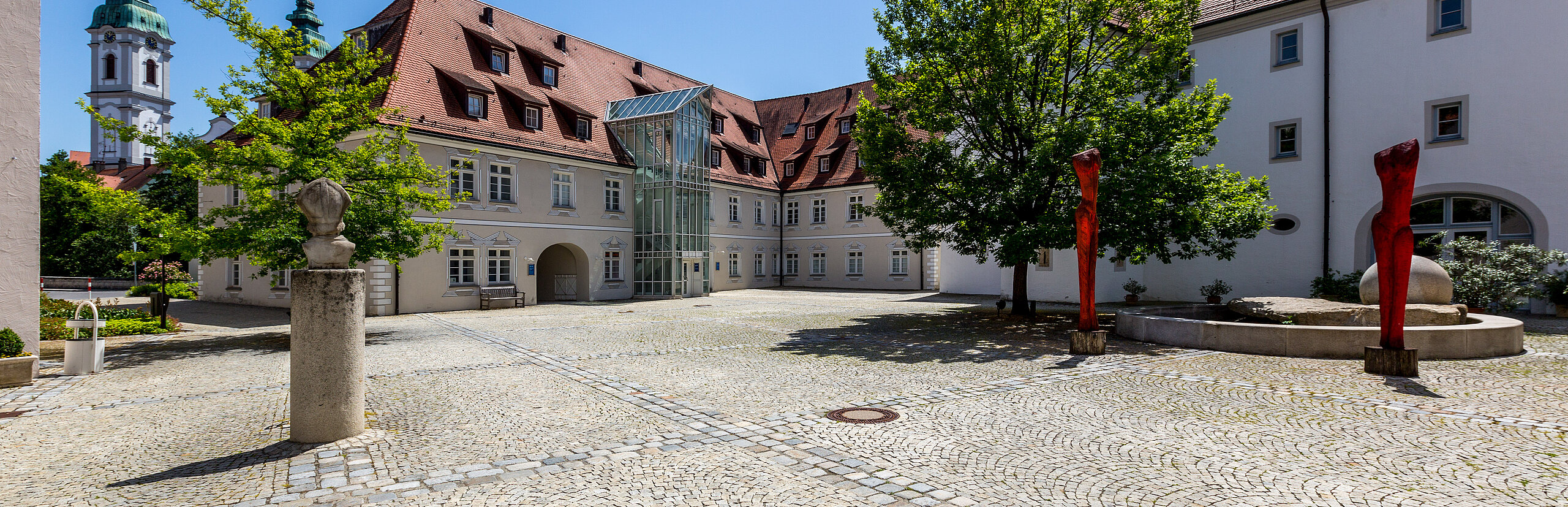 Gepflasterter Innenhof der Klinik im Hofgarten in Bad Waldsee, umgeben von historischen Gebäuden mit roten Dächern, Bäumen und blauem Himmel im Hintergrund